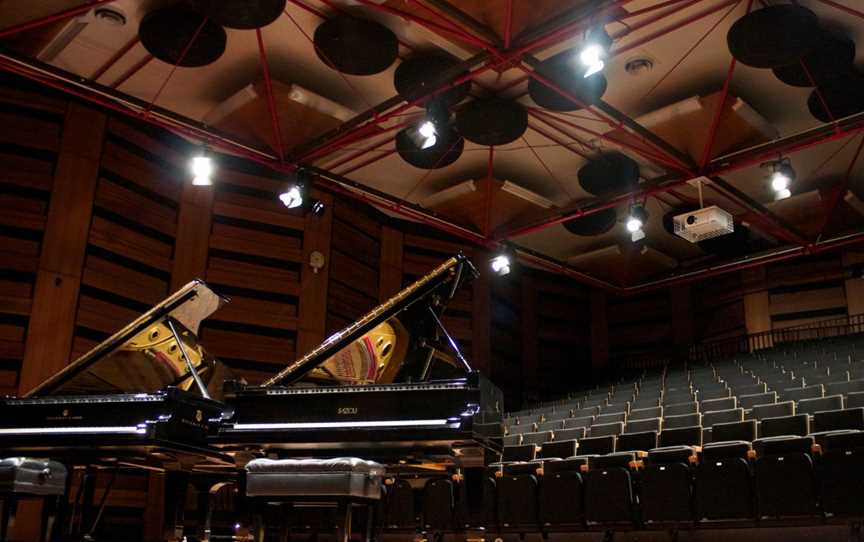 Two shiny black pianos have their lids opened and are standing side by side, in front of the ranked seating in a performance space.