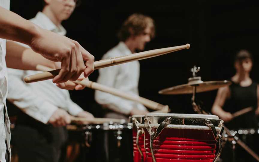 Foreground shows a pair of hand holding wooden drumsticks which are hitting the head of a pair of bright red drums.